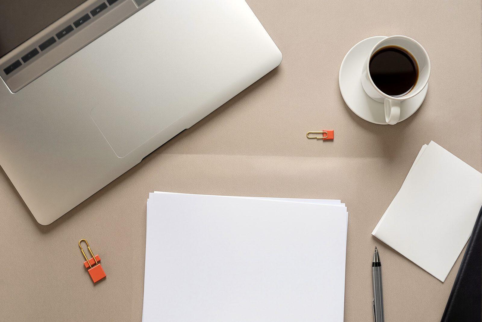 A desk seen from above with a closed laptop, a cup of coffee, blank paper and one vermilion paper clip