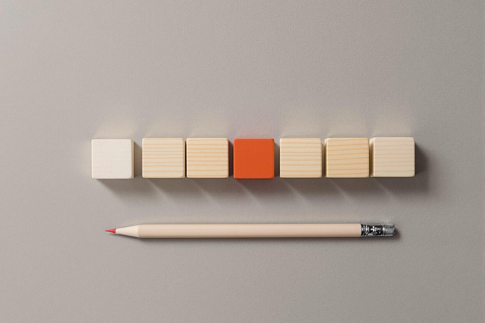 Seven small wooden blocks in a row on grey paper, the fifth painted vermilion, a pencil beside them