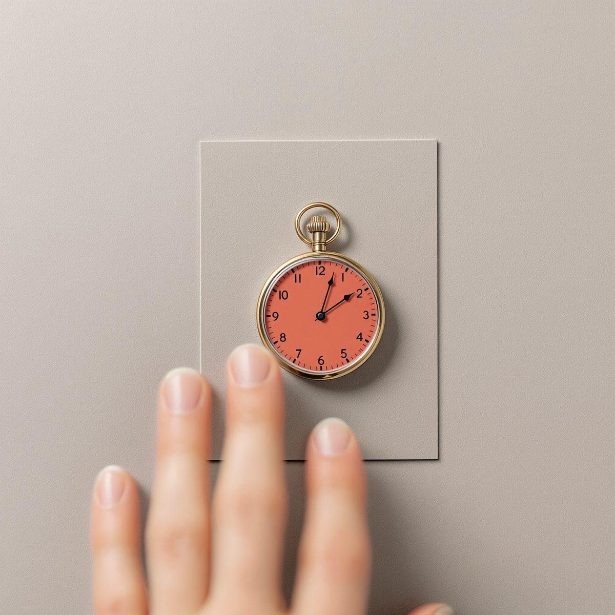 A stopwatch with a vermilion face lying on grey card, a hand reaching for it